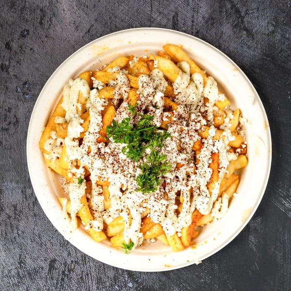 Golden french fries topped with white sauce, crumbled cheese, sumac, and parsley in a bowl.