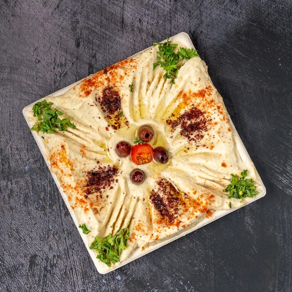 Square plate of hummus, garnished with olive oil, paprika, parsley, olives, and a cherry tomato.