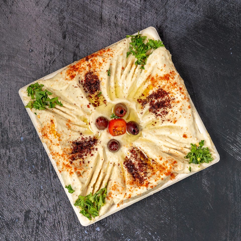 Square plate of hummus, garnished with olive oil, paprika, parsley, olives, and a cherry tomato.
