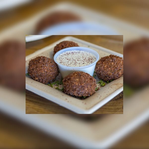 Four falafel balls surround a small cup of white sauce sprinkled with spices on a square plate.